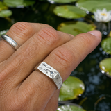 A close-up of a hand wearing a silver band ring with engraved frog illustrations, set against a background of green lily pads and water.