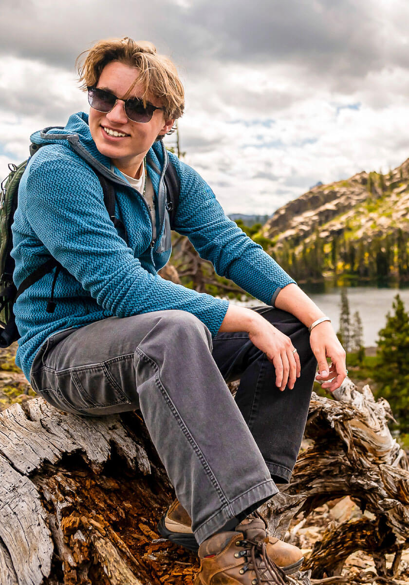 Person sitting on a log with a scenic background of mountains and water.