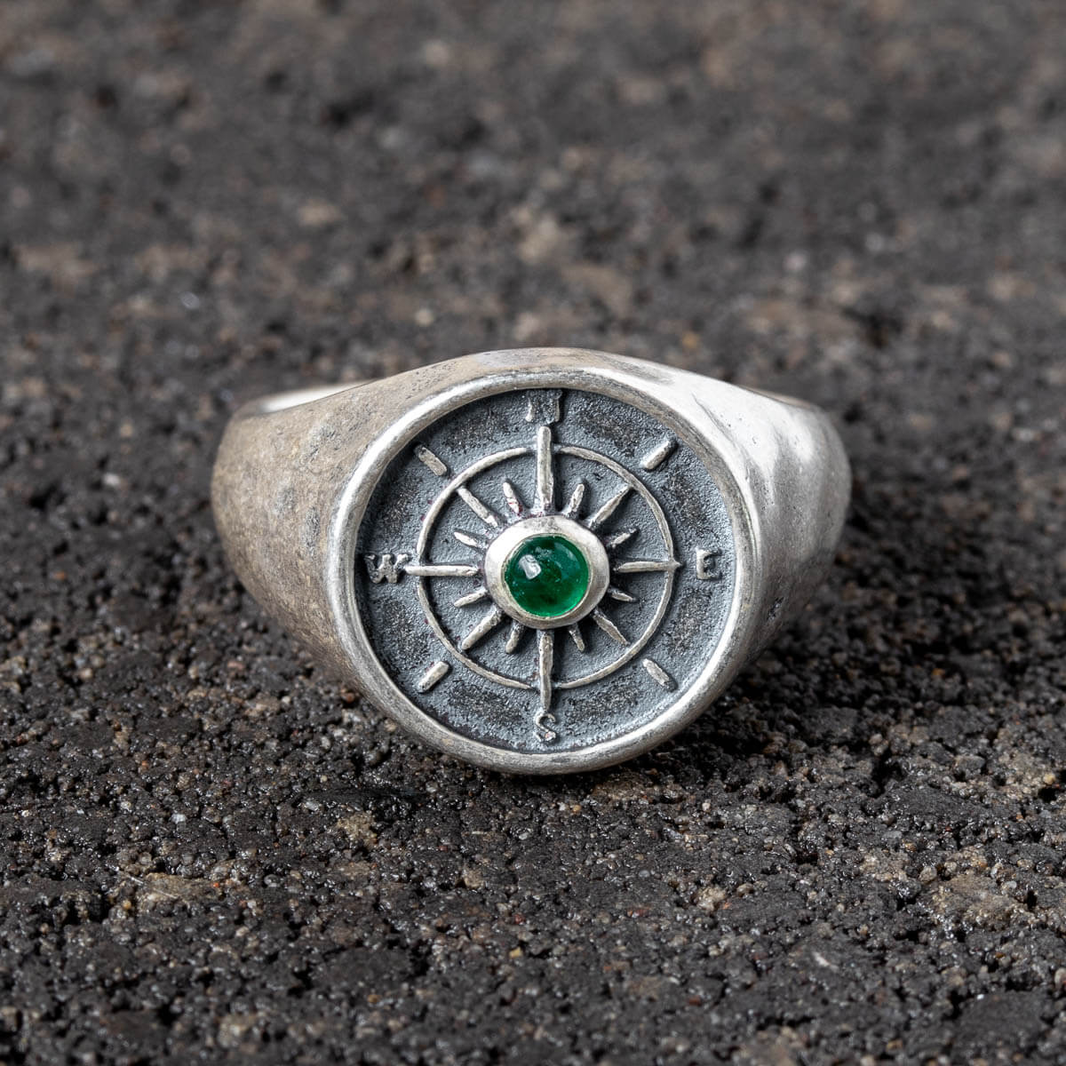 Silver ring with a compass design and emerald gemstone on a dark textured surface