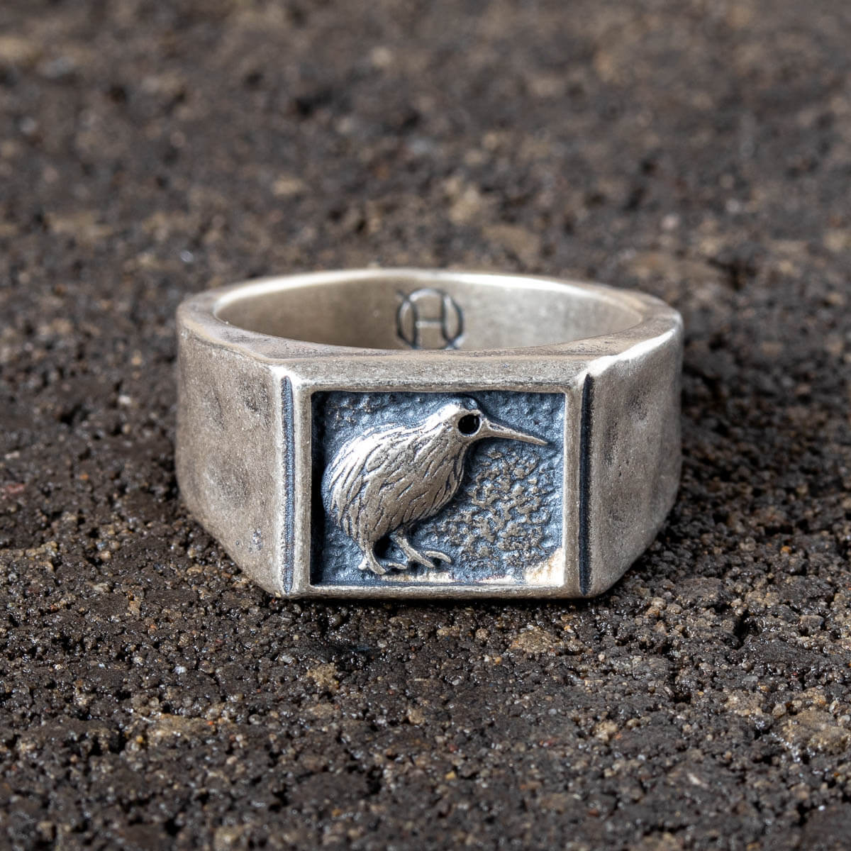 Silver ring with a kiwi bird emblem on a dark textured surface