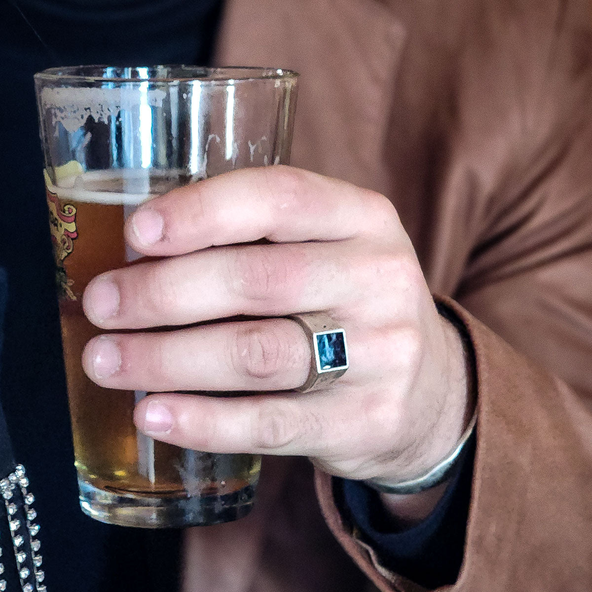 Hand holding a glass of beer with a brown jacket wearing silver pietersite ring