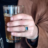 Hand holding a glass of beer with a brown jacket wearing silver pietersite ring
