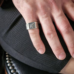 Hand wearing a silver ring with a detailed design on a dark textured surface