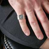 Hand wearing a silver ring with a detailed design on a dark textured surface