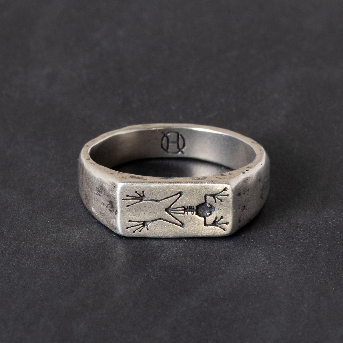 A silver signet ring with engraved frog skeleton on a dark textured background.