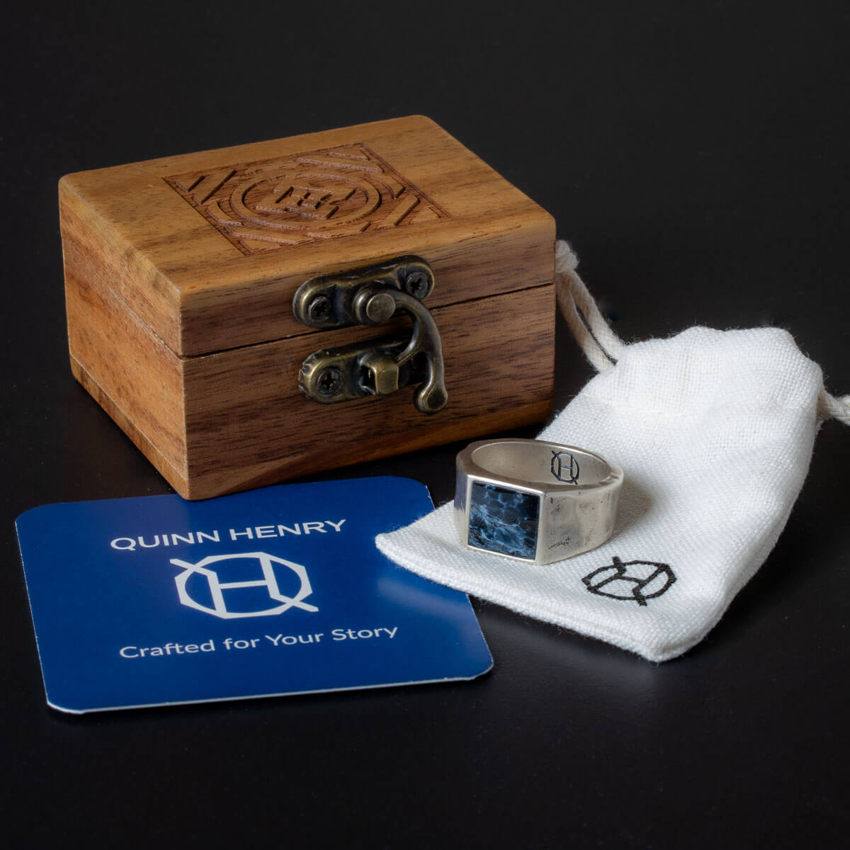 Wooden box with a silver ring on a white cloth, branded 'Quinn Henry', on a dark background.