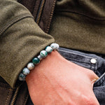 Close-up of a hand wearing a bracelet with green beads, against a background of a jacket and jeans.