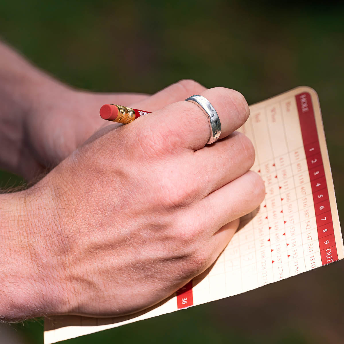 Hand with a ring holding a red pencil over a golf score card on a blurred green background