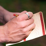 Hand with a ring holding a red pencil over a golf score card on a blurred green background