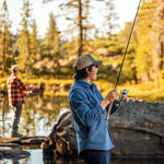 Two people fishing by a lake with trees in the background