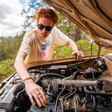 Person working on a car engine outdoors with trees in the background