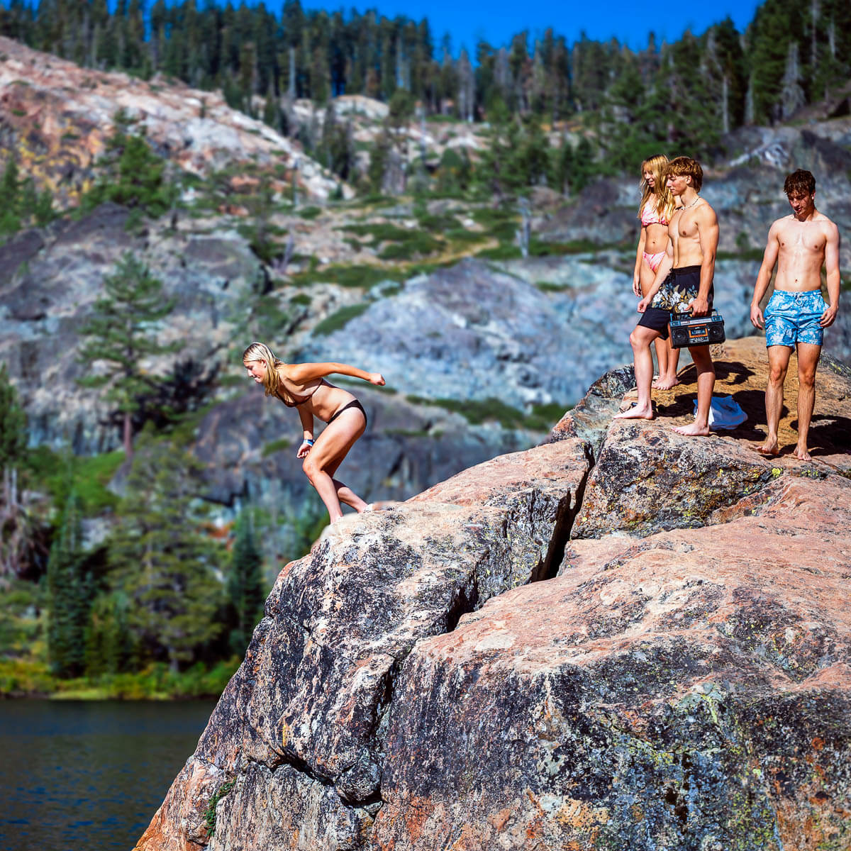 woman jumping off rock outcropping into lake as friends watch