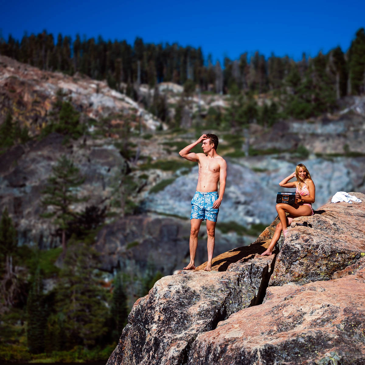 Two people on a rocky outcrop with a scenic background of trees and mountains.